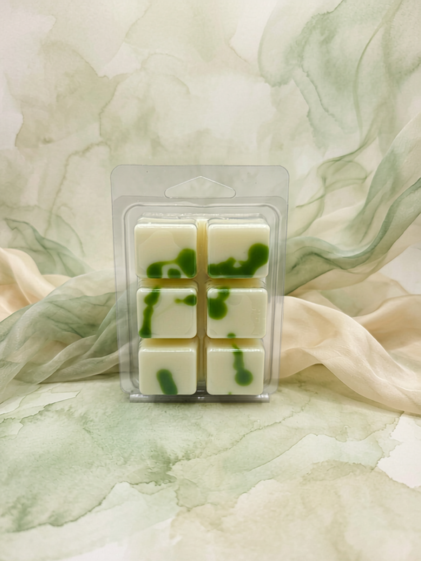 Six square white wax melts with green marbled swirls, packaged in a clear plastic clamshell blister pack, set against a soft watercolor-style green and cream background with draped fabric.