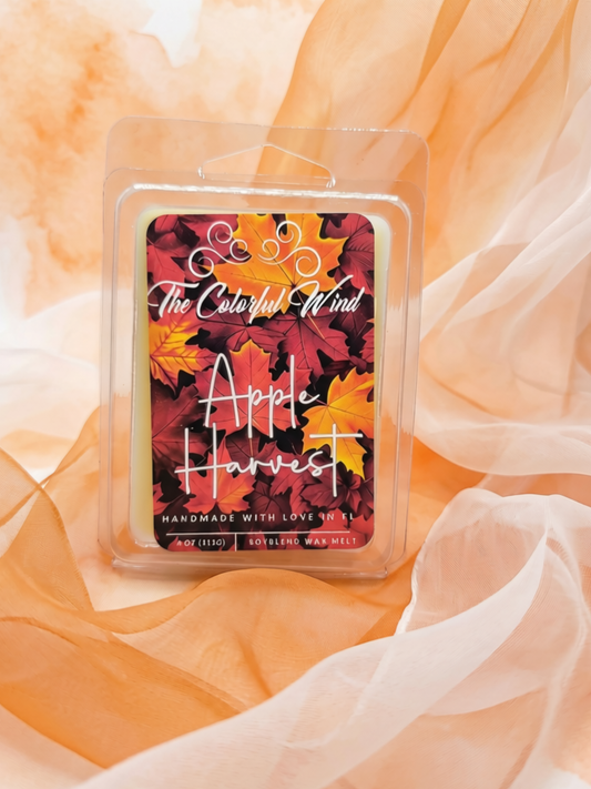 An apple harvest scented wax melt in a clear clamshell package, featuring a label with colorful autumn leaves in red, orange, and yellow, and white script text reading 'The Colorful Wind' and 'Apple Harvest'. The package is set against a soft, draped peach fabric background.