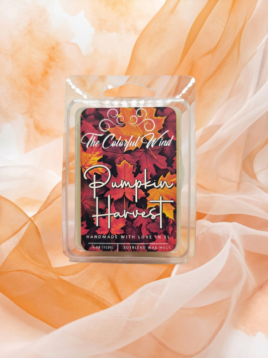 A packaged wax melt product labeled 'The Colorful Wind' and 'Pumpkin Harvest' sits centered on draped peach fabric. The front label features a floral autumn design with red, orange, and yellow leaves, and the wax melt container is clear plastic.